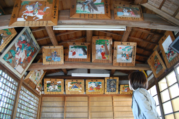 木造の建物の中、一人の女性が天井に飾られたたくさんの絵画を見上げている写真。